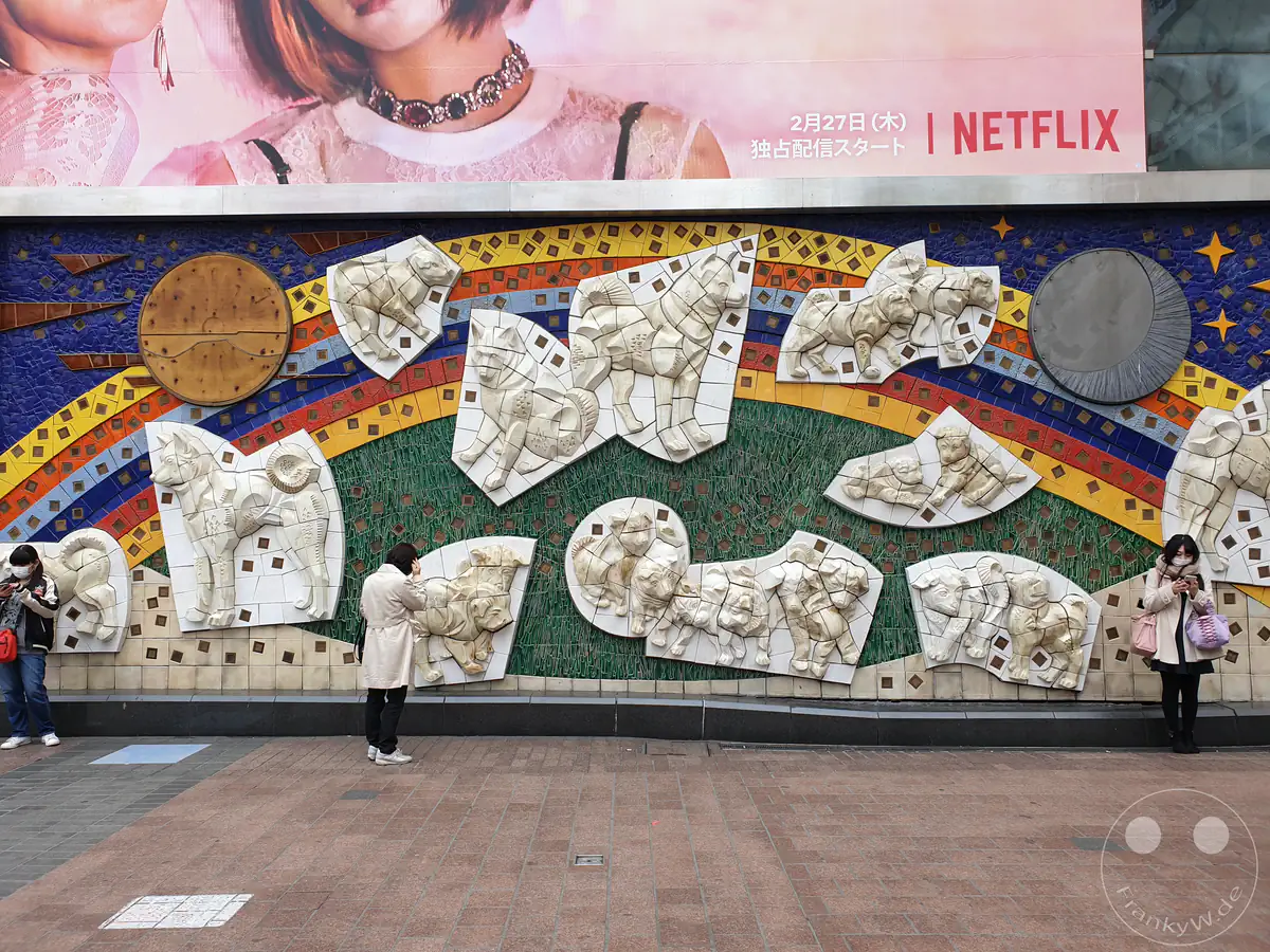 Das berühmte Keramik-Mosaik-Relief „Shibuya-Welt der Hunde“ des Künstlers Yoshiko Miyashita an der Außenwand des Shibuya-Bahnhofs zeigt eine Darstellung von Akita-Inu-Hunden unter einer großen Netflix-Plakatwerbung mit Passanten im Stadtteil Shibuya in Tokio, Japan.