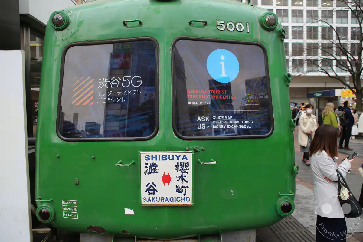 Die historische grüne Front eines alten Zugwaggons der Tokyu-5000-Serie, bekannt als Grüner Frosch (Aogaeru), dient als Touristeninformationszentrum auf dem Hachiko-Platz vor dem Bahnhof Shibuya mit Passanten im Stadtteil Shibuya in Tokio, Japan.