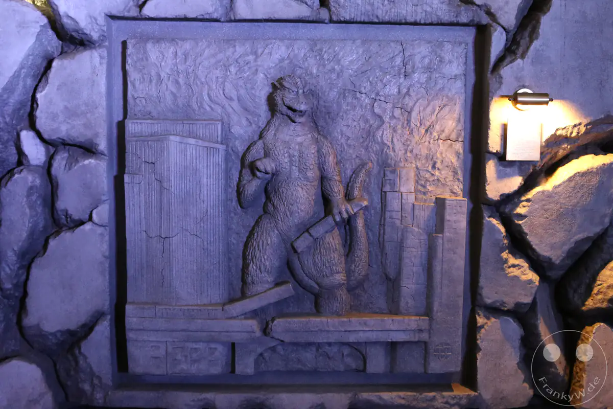 Ein kunstvolles, bläulich beleuchtetes Steinrelief an einer Wand zeigt das ikonische Filmmonster Godzilla inmitten von zerstörten Brückenteilen und Hochhäusern in der Nähe des Hotel Gracery im Stadtteil Shinjuku in Tokio, Japan.