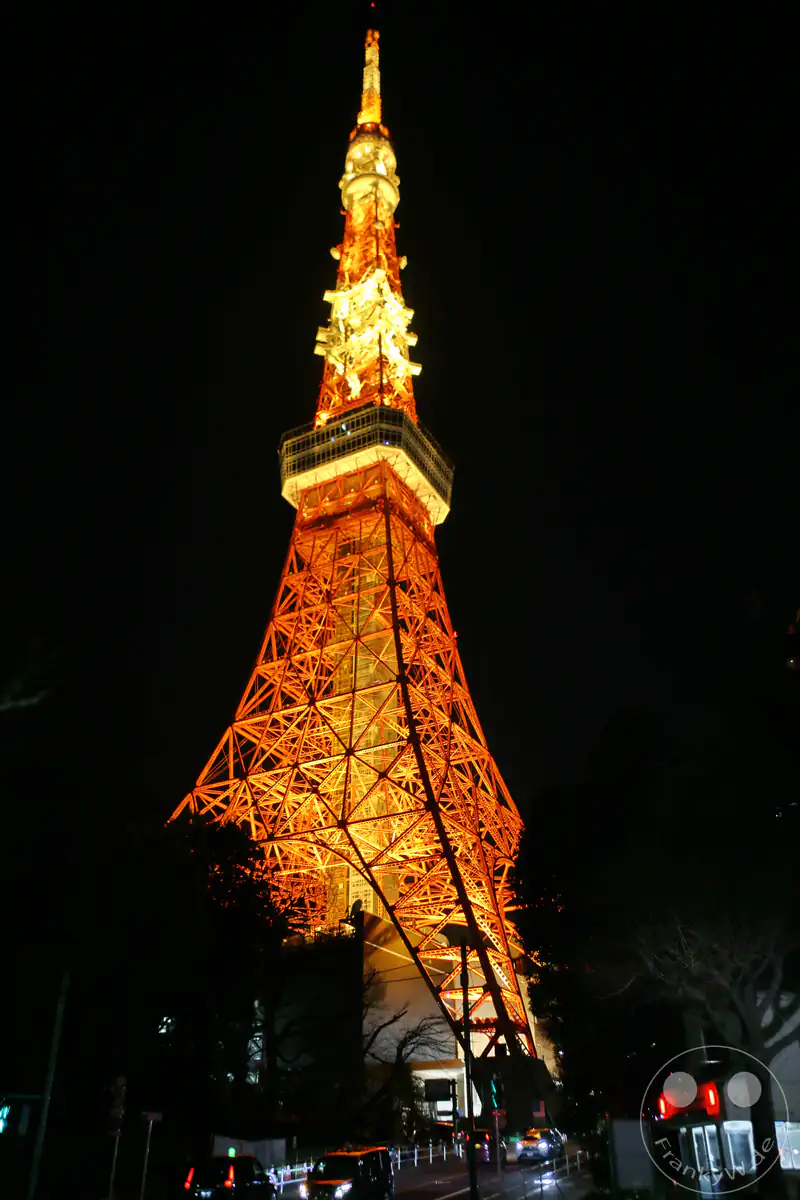 Der ikonische Tokyo Tower erstrahlt bei Nacht in einer intensiv orange-roten Festbeleuchtung und ragt als leuchtendes Wahrzeichen hinter einer nächtlichen Straße mit Autos in den dunklen Himmel von Tokio, Japan.