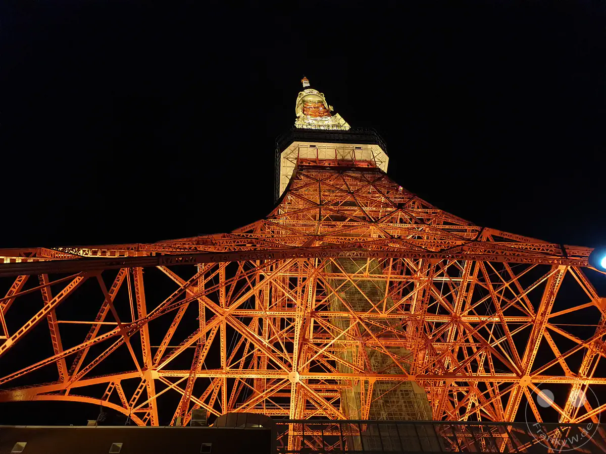Eine direkte Sicht von unten auf den festlich illuminierten Tokyo Tower bei Nacht, die das komplexe Geflecht der orange-roten Stahlträger und die hell erleuchtete Hauptplattform vor dem schwarzen Himmel in Tokio, Japan, detailreich einfängt.