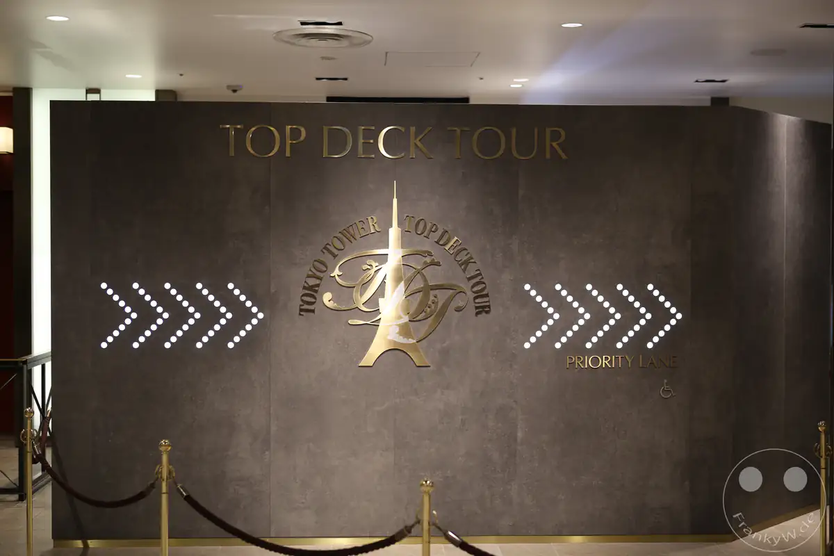 Eine graue Trennwand im Innenbereich des Tokyo Tower markiert den Zugang zur Top Deck Tour mit einem goldenen Logo, dem Schriftzug Priority Lane und leuchtenden LED-Pfeilen zur Orientierung für Besucher in Tokio, Japan.