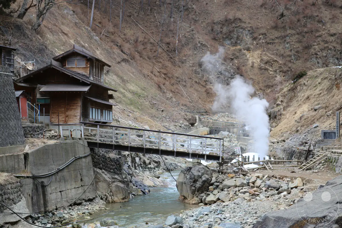 Historisches Korakukan-Ryokan aus Holz neben einer Fußgängerbrücke über einen steinigen Gebirgsfluss mit einer dampfenden Geysir-Quelle im Jigokudani-Tal in Yamanouchi in der Präfektur Nagano, Japan.