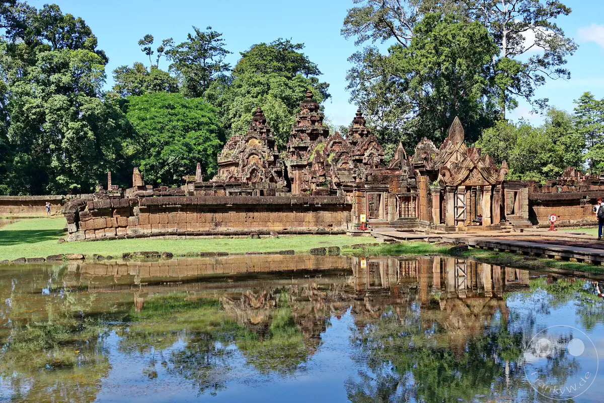 Die Außenansicht des historischen Banteay-Srei-Tempels aus rotem Sandstein, dessen reich verzierte Steintürme und Umfassungsmauern sich in einem Wasserbecken im Vordergrund spiegeln.