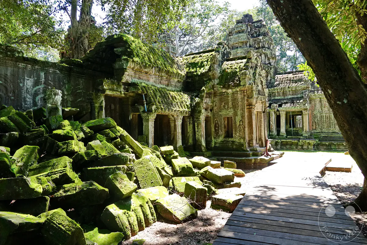 Ein großer Haufen eingestürzter, dicht mit grünem Moos bewachsener Steinblöcke neben einem hölzernen Besucherweg vor den historischen, verfallenen Tempelstrukturen von Ta Prohm in Angkor.