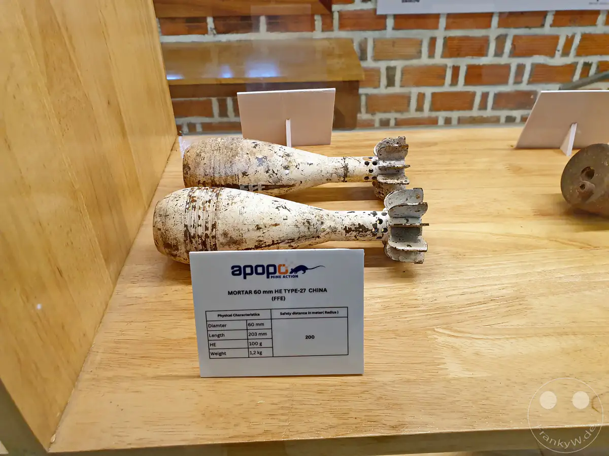 Zwei historische, entschärfte chinesische 60-mm-Mörsergranaten vom Typ 27 (FFE - Free From Explosives) auf einem Holztisch im APOPO-Besucherzentrum mit einem Informationsschild zu den technischen Daten