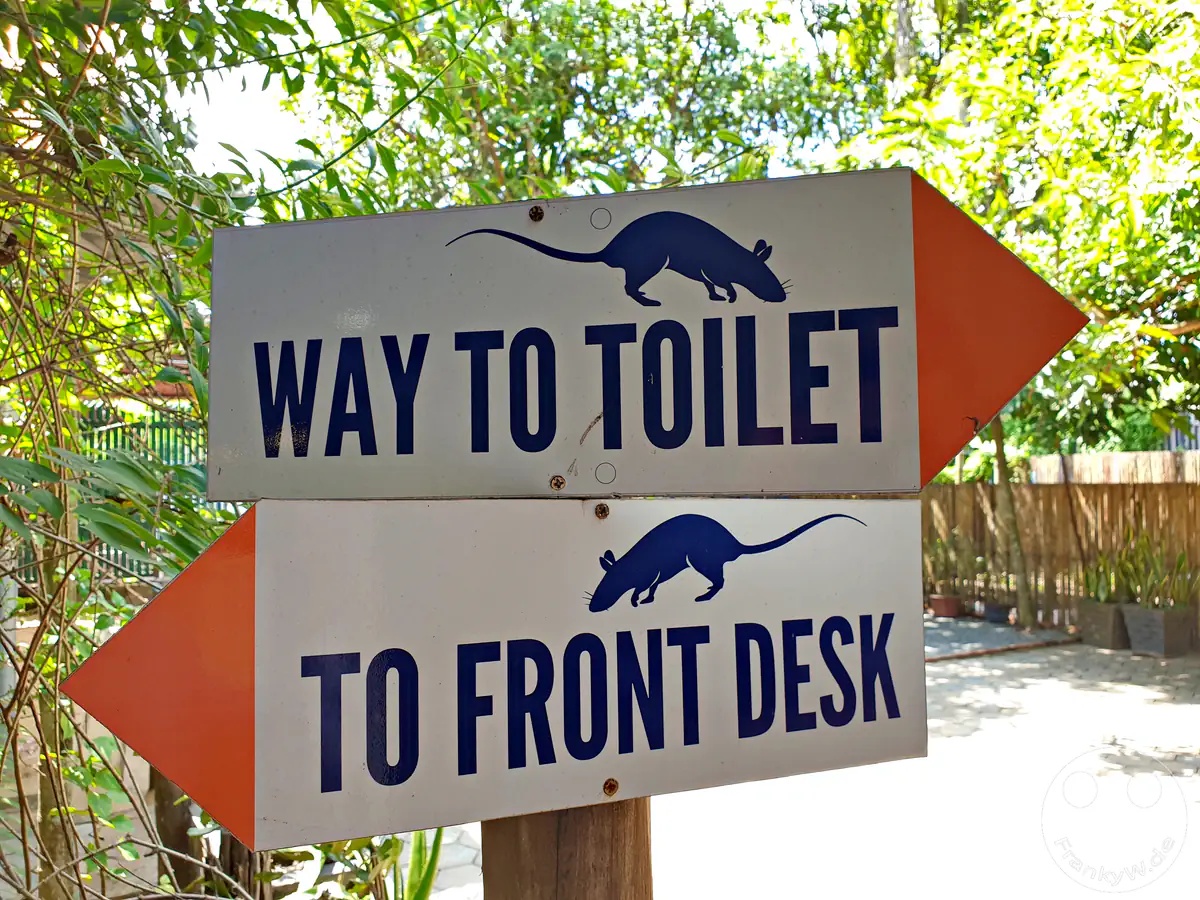 Ein hölzerner Wegweiser im APOPO-Besucherzentrum mit zwei pfeilförmigen Schildern, die mit Ratten-Silhouetten verziert sind und die Richtungen „WAY TO TOILET“ nach rechts sowie „TO FRONT DESK“ nach links anzeigen.