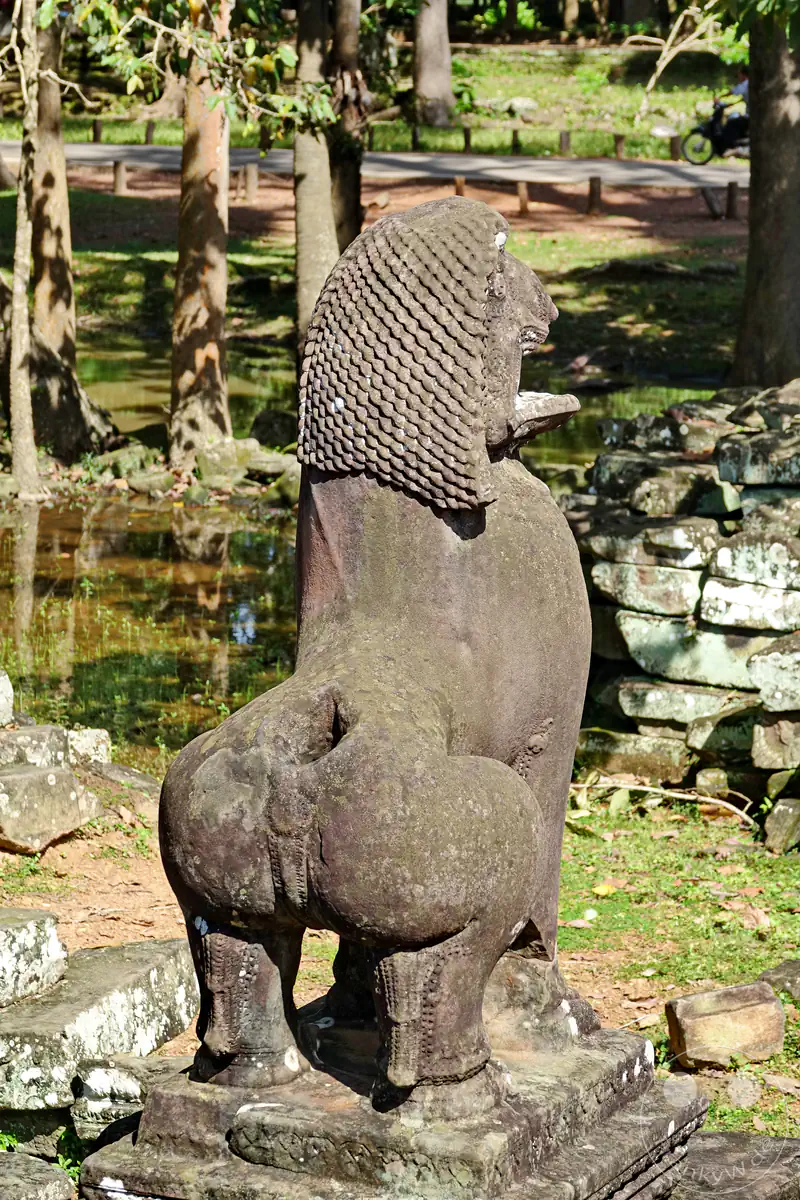 Eine historische steinerne Löwenstatue (Singh) als traditionelle Wächterfigur an den Tempelruinen von Angkor in Kambodscha vor Bäumen und einem Wasserlauf.