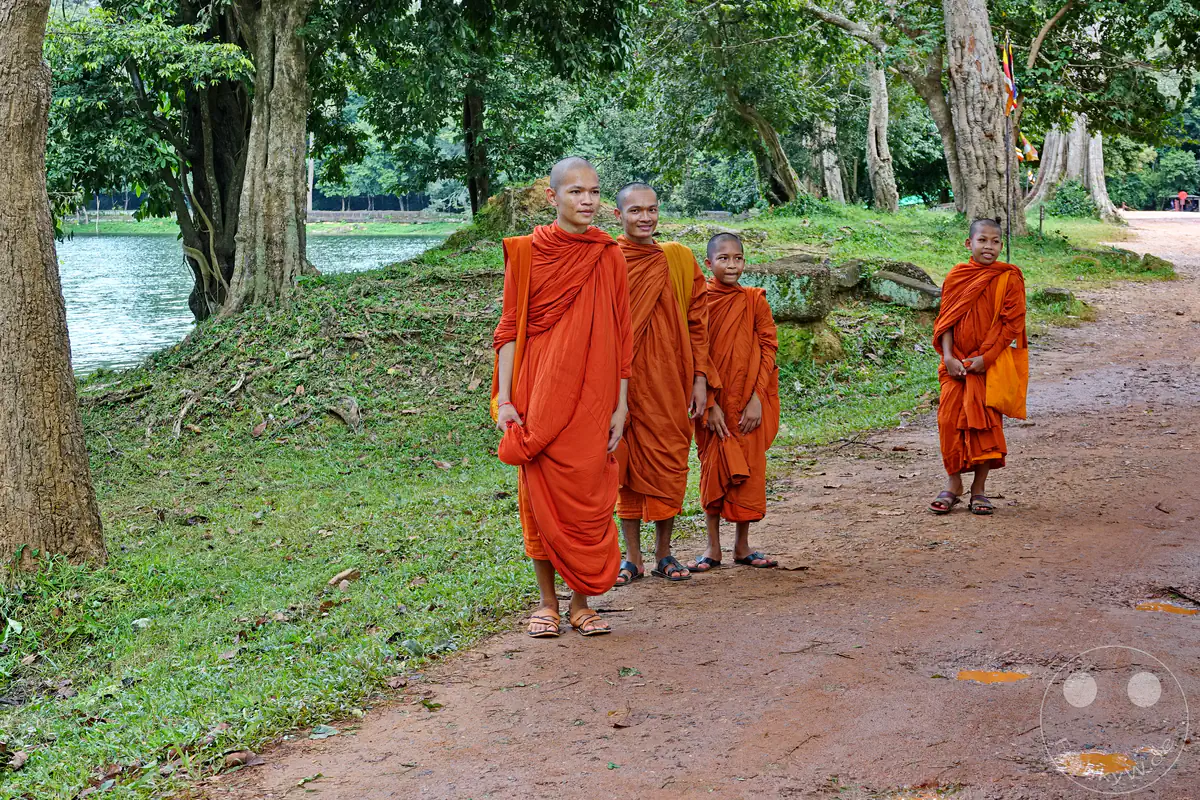 Eine Gruppe von vier jungen buddhistischen Mönchen und Novizen in traditionellen orangefarbenen Roben steht lächelnd auf einem unbefestigten Erdweg nahe einem Gewässer und alten Steinruinen im Tempelbezirk von Angkor.