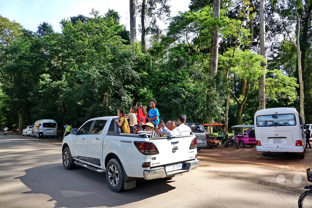 Ein weißer Pickup-Truck mit Passagieren auf der Ladefläche fährt auf einer Straße nahe den historischen Tempelanlagen von Angkor in Kambodscha, umgeben von parkenden Fahrzeugen und dichtem Wald