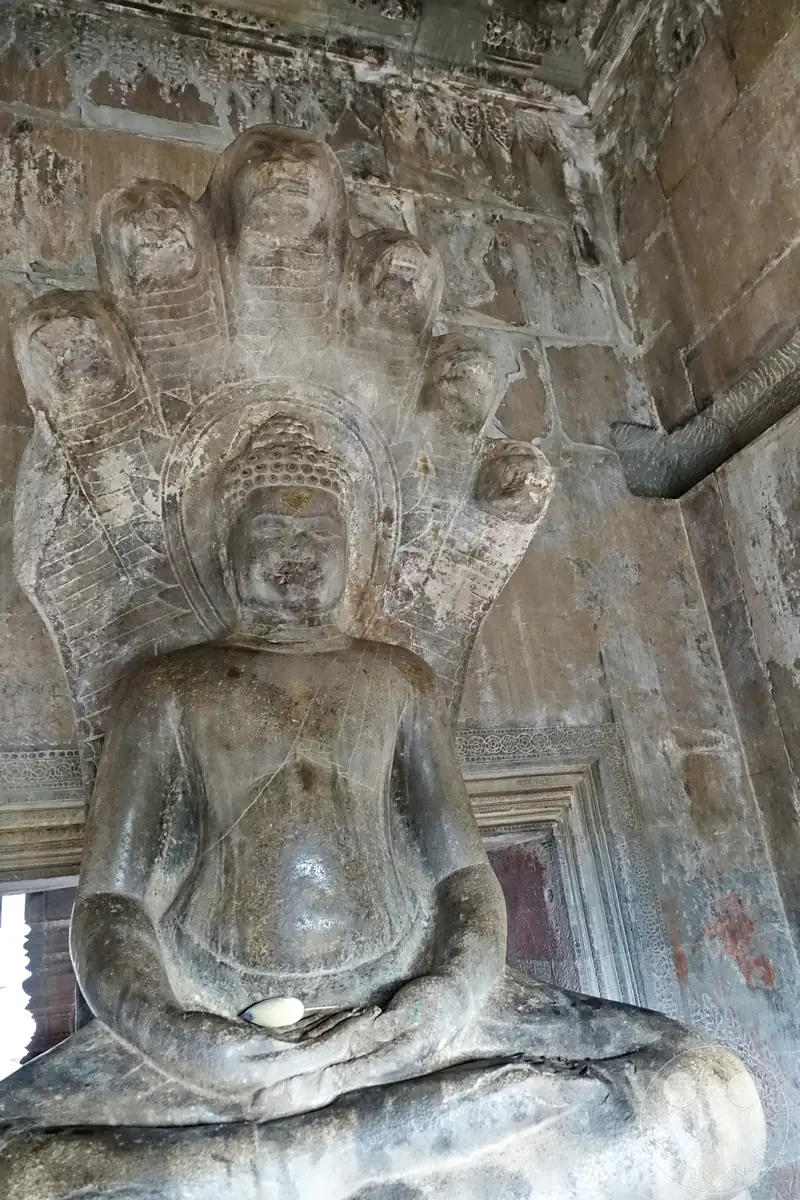 Eine historische steinerne Buddha-Statue im Dhyana-Mudra-Gestus der Meditation, sitzend auf den Windungen der Schlage Mucalinda, deren vielköpfiger Naga-Schirm über dem Kopf des Erleuchteten in einer Tempelhalle von Angkor Wat emporragt.