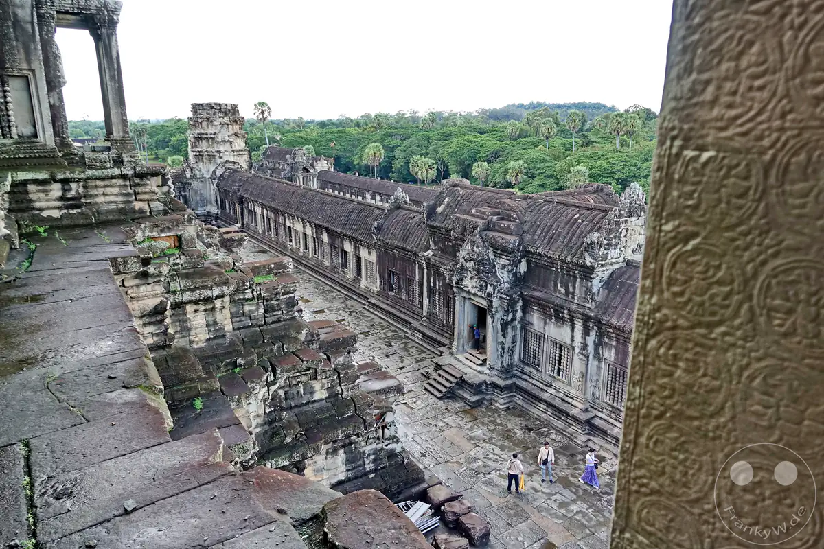 Der Blick von einer oberen Ebene des Angkor-Wat-Tempels hinab auf die langen, historischen Galeriebauten aus Stein und einige Besucher im Innenhof vor der Kulisse des angrenzenden Dschungelwaldes.