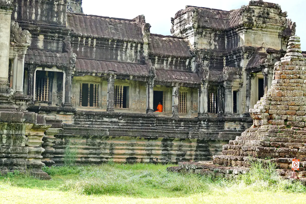 Ein buddhistischer Mönch in einer leuchtend orangefarbenen Robe sitzt in einem schattigen Fenster der historischen, steinernen Galeriebauten von Angkor Wat, flankiert von verzierten Steinsäulen und einem kleinen Stupa im Vordergrund.