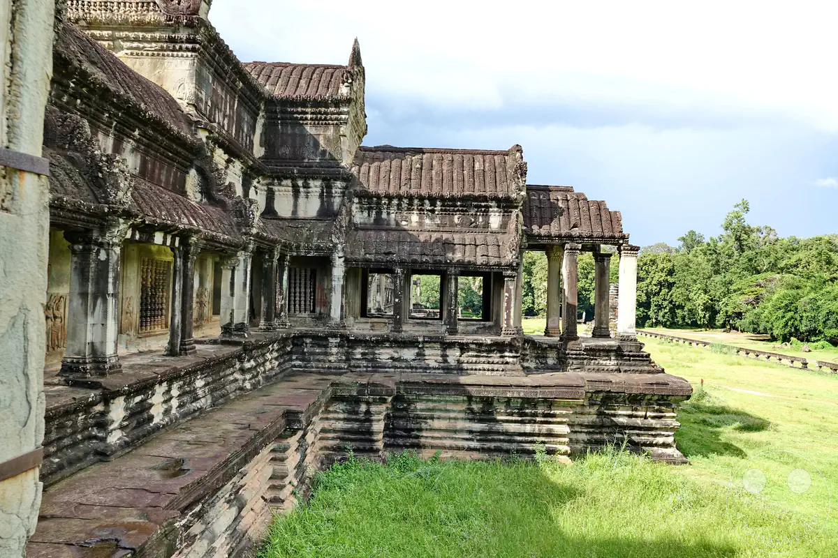 Die perspektivische Außenansicht der historischen, terrassenförmig abgestuften Galeriebauten in Angkor Wat mit von verzierten Steinpfeilern getragenen Hallen über einer grünen Rasenfläche vor dichtem Wald.