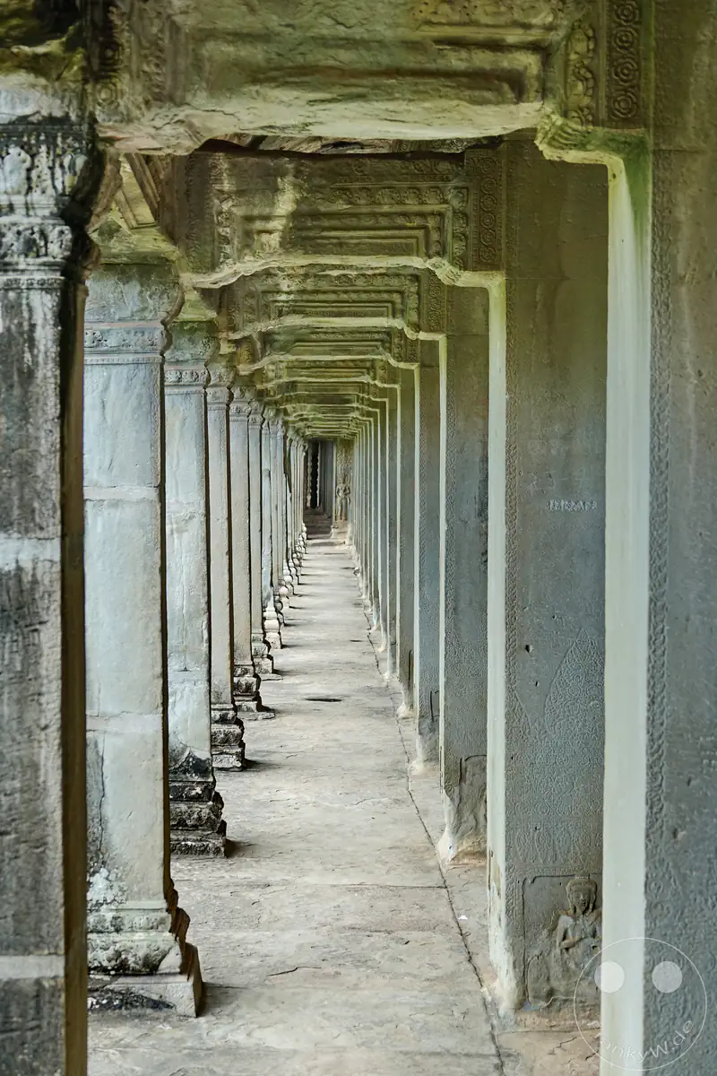 Der Blick durch einen langen, historischen Galeriegang in der Tempelanlage Angkor Wat mit einer tiefen Flucht aus verzierten Steinsäulen und Durchgängen.