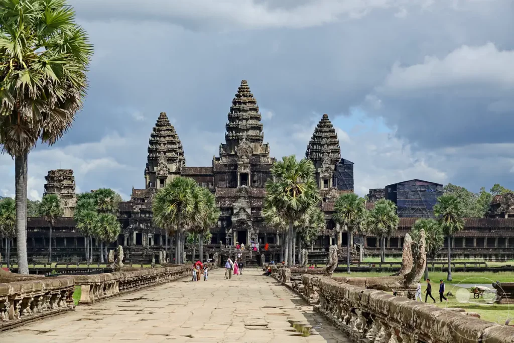 Der Blick entlang des breiten, historischen Steinstegs auf die berühmte Hauptfassade von Angkor Wat mit ihren drei markanten lutosförmigen Türmen und flankierenden Palmen unter bewölktem Himmel.