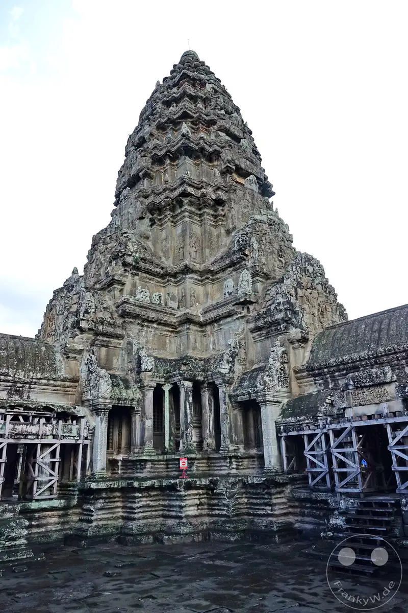 Der monumentale, zentrale Steinturm von Angkor Wat erhebt sich steil über einem historischen Innenhof mit hölzernen Stützkonstruktionen und Treppenaufgängen an den Seiten unter einem hellen Himmel.