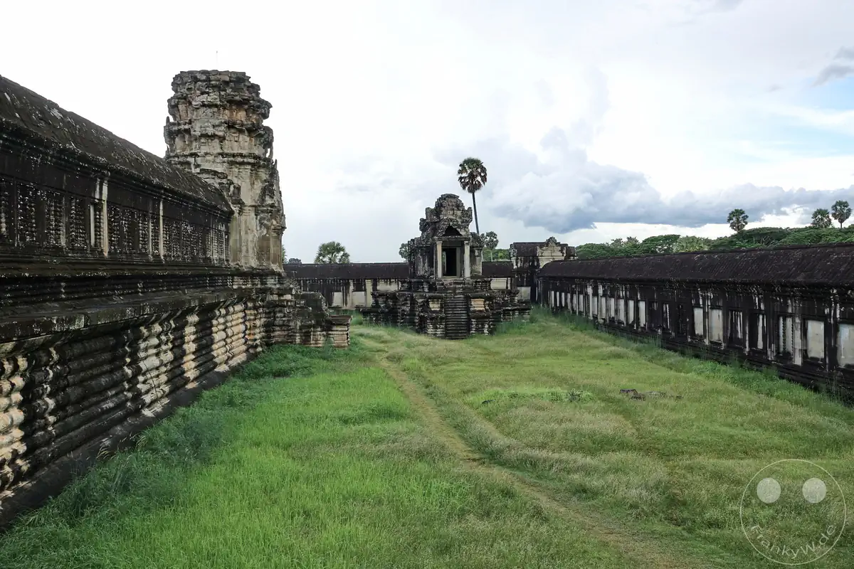 Ein weitläufiger, historischer Innenhof der Tempelanlage Angkor Wat mit einer grünen Grasfläche, einem freistehenden Steinturm in der Mitte und langen Galeriebauten an den Außenseiten unter bewölktem Himmel.