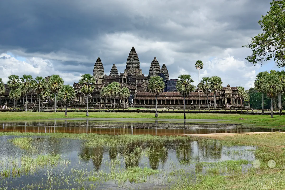 Die weite Außenansicht der historischen Tempelanlage Angkor Wat in Kambodscha mit ihren charakteristischen lofusförmigen Steintürmen, die sich zusammen mit den umliegenden Palmen in einem flachen Seerosenbecken im Vordergrund spiegeln.
