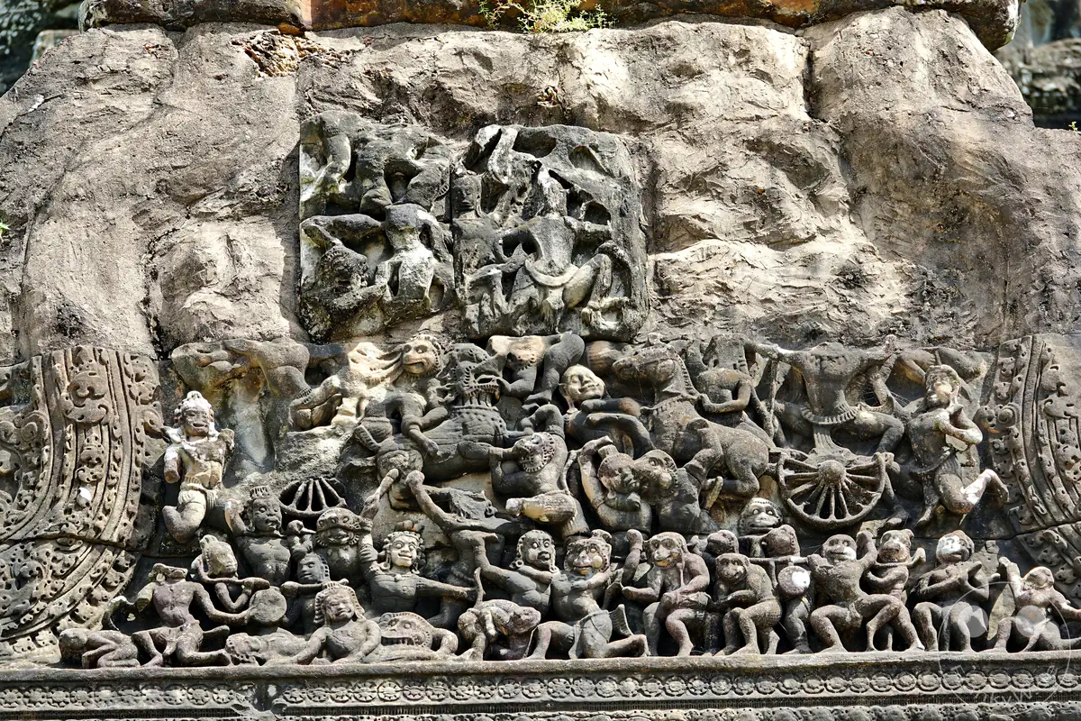 Ein detailreiches historisches Steinrelief an einer Tempelmauer in Angkor Wat mit komplexen Darstellungen von Figuren aus der hinduistischen Mythologie, Affenkriegern, Wagen und einer Schlachtenszene.