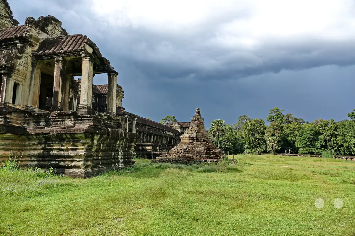 Ein freistehender, kleiner Steinstupa auf einer weiten grünen Wiese neben den historischen Galeriebauten von Angkor Wat vor dichten Dschungelbäumen unter einem dunkel bewölkten Himmel.