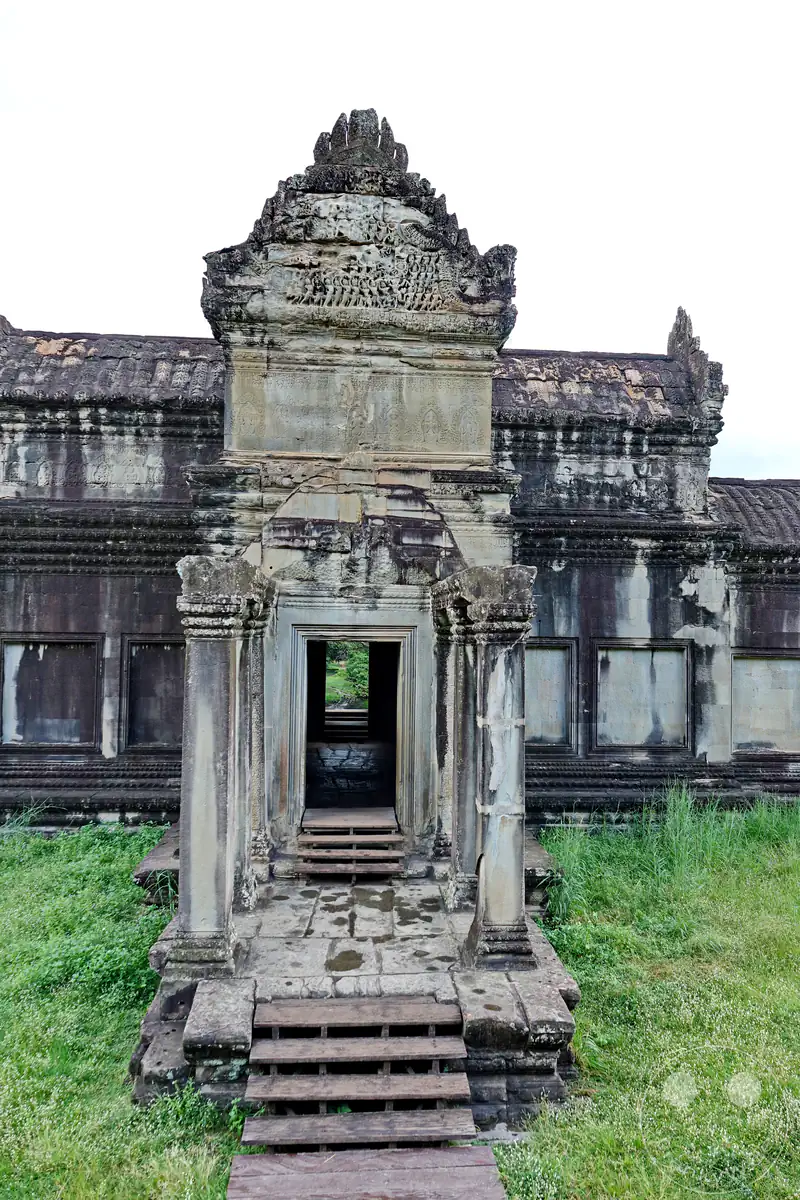 Ein historischer hölzerner Treppenaufgang führt zu einem verzierten Steintor mit einem reich gemeißelten Giebelrelief an den Galeriebauten von Angkor Wat vor einer grünen Grasfläche.