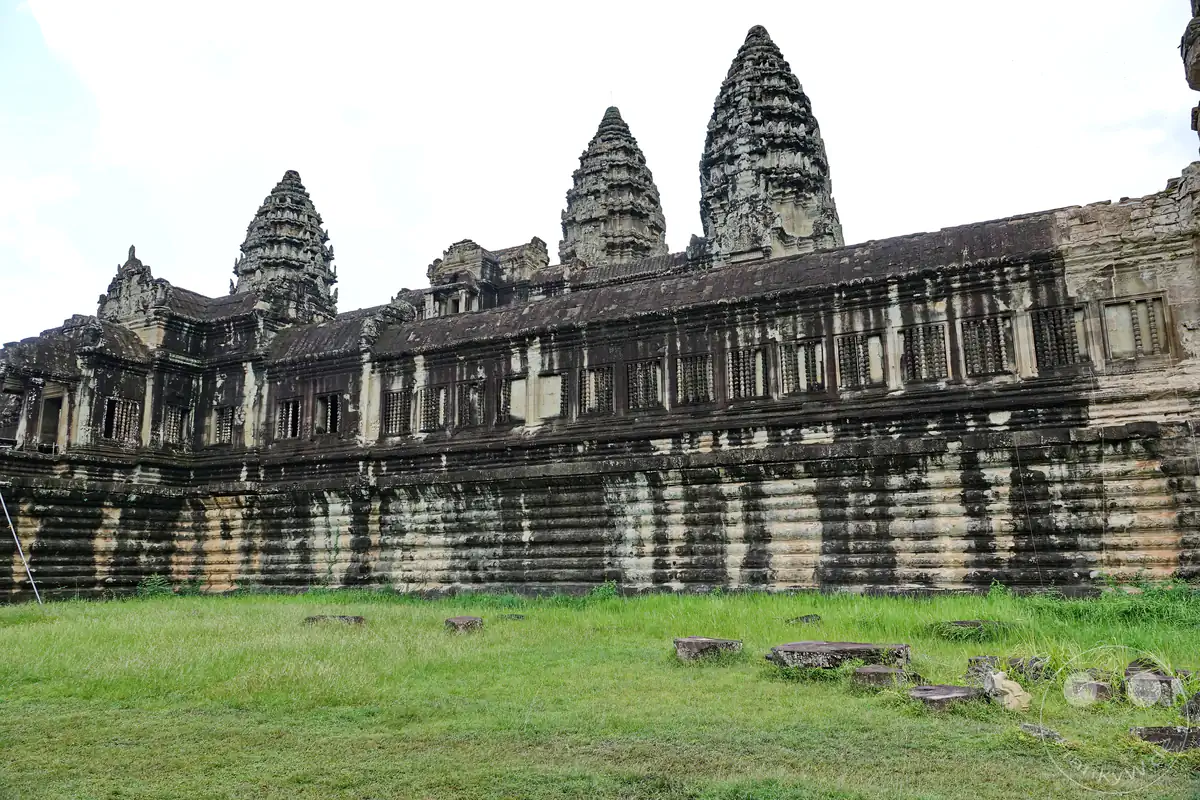 Die weite Außenansicht der historischen, langgestreckten Galeriebauten aus Stein in Angkor Wat, über deren Mauern sich die charakteristischen, lotusförmigen Tempeltürme hinter einer grünen Rasenfläche erheben.