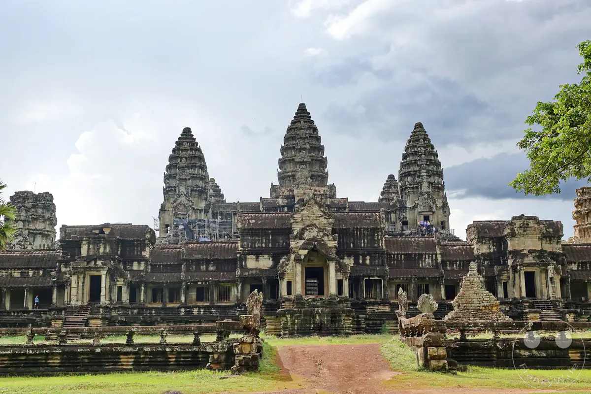 Die monumentale Frontansicht des historischen Angkor-Wat-Tempels mit seinen drei markanten, lotusförmigen Haupttürmen über den weitläufigen Galeriebauten, vor denen ein unbefestigter roter Erdweg und eine grüne Rasenfläche unter bewölktem Himmel liegen.