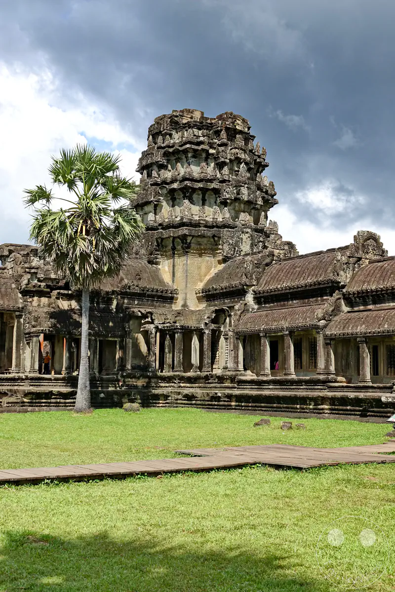Ein markanter historischer Steinturm und die angrenzenden Galeriebauten von Angkor Wat hinter einer grünen Rasenfläche mit einer Palme und einem Holzsteg unter bewölktem Himmel.