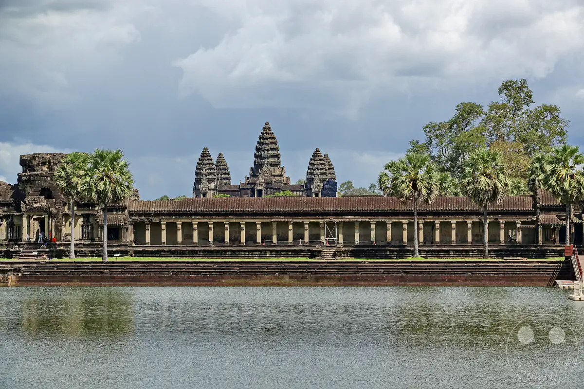 Die markante Außenansicht des historischen Angkor-Wat-Tempels in Kambodscha mit seinen charakteristischen lutosförmigen Türmen hinter der langen Galerie und Palmen, die sich im großen Wassergraben im Vordergrund spiegeln.