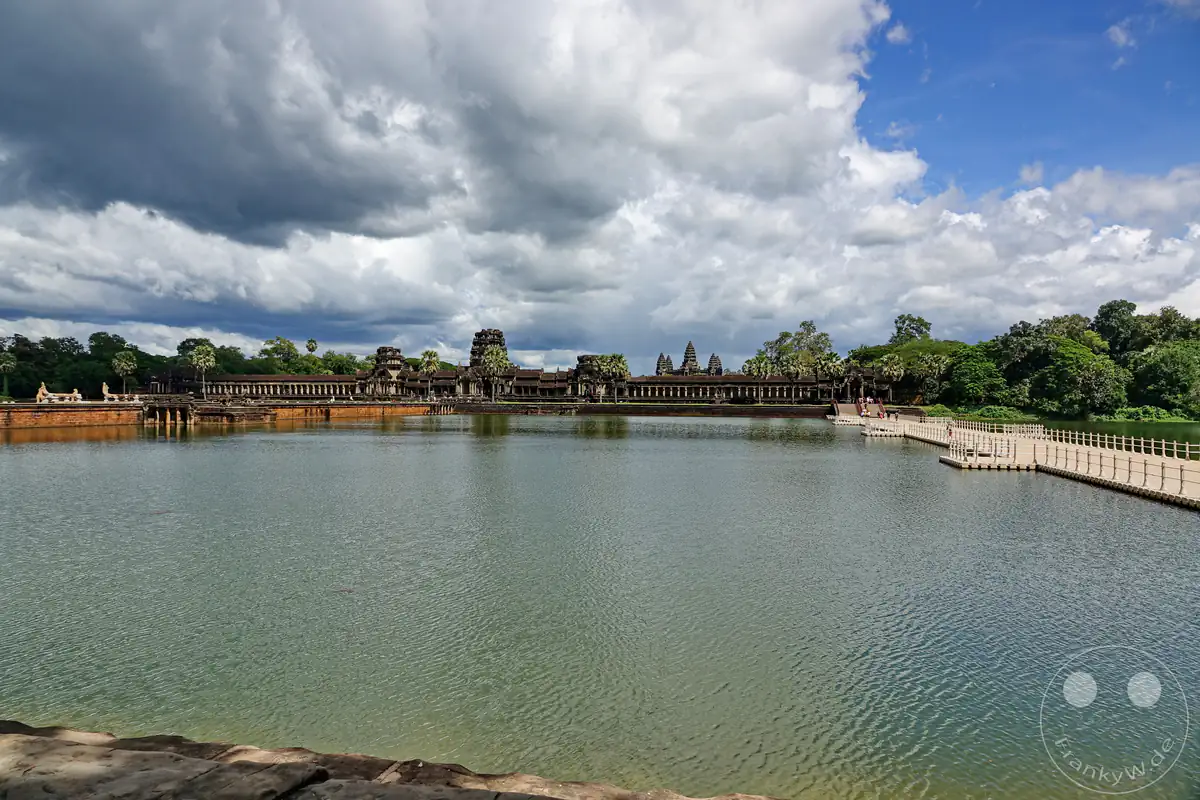 Die weite Panorama-Außenansicht der historischen Tempelanlage Angkor Wat mit dem breiten Wassergraben im Vordergrund und einer modernen, schwimmenden Fußgängerbrücke auf der rechten Seite unter einem dramatisch bewölkten Himmel.