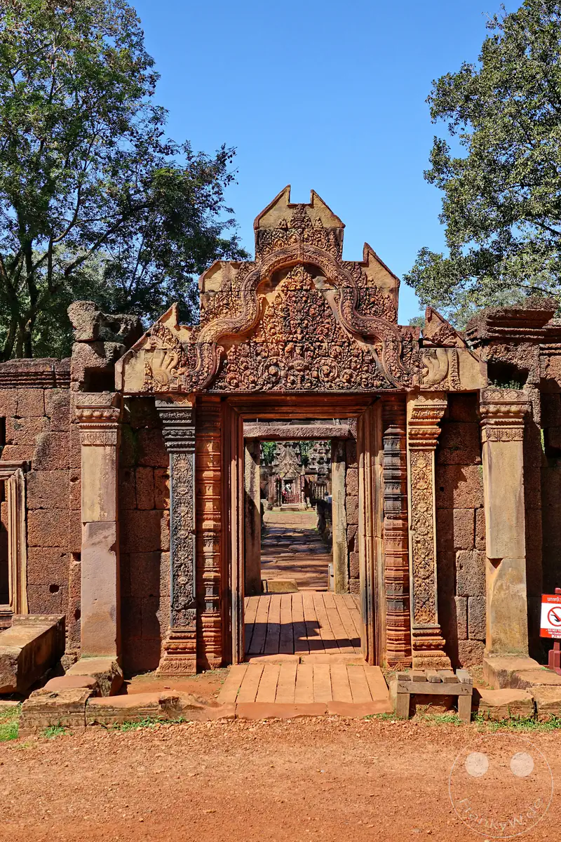 Der Blick durch ein reich verziertes, historisches Steintor aus rotem Sandstein im Banteay-Srei-Tempel, das detailreiche Giebelreliefs und kunstvolle Steinmetzarbeiten vor einem klaren blauen Himmel zeigt.