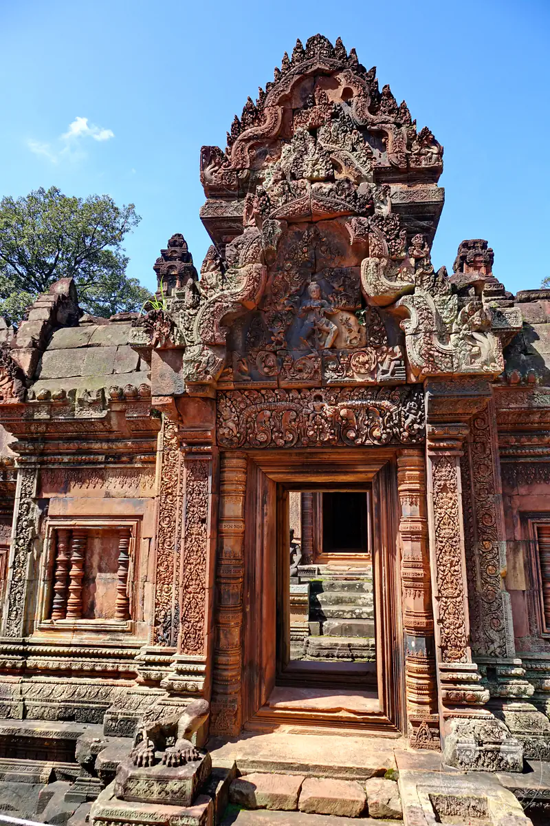 Ein detailreich verzierter Toreingang aus rotem Sandstein im Banteay-Srei-Tempel mit einem kunstvollen Giebelrelief, das eine hinduistische Gottheit darstellt, flankiert von feinen Steinmetzarbeiten unter blauem Himmel.