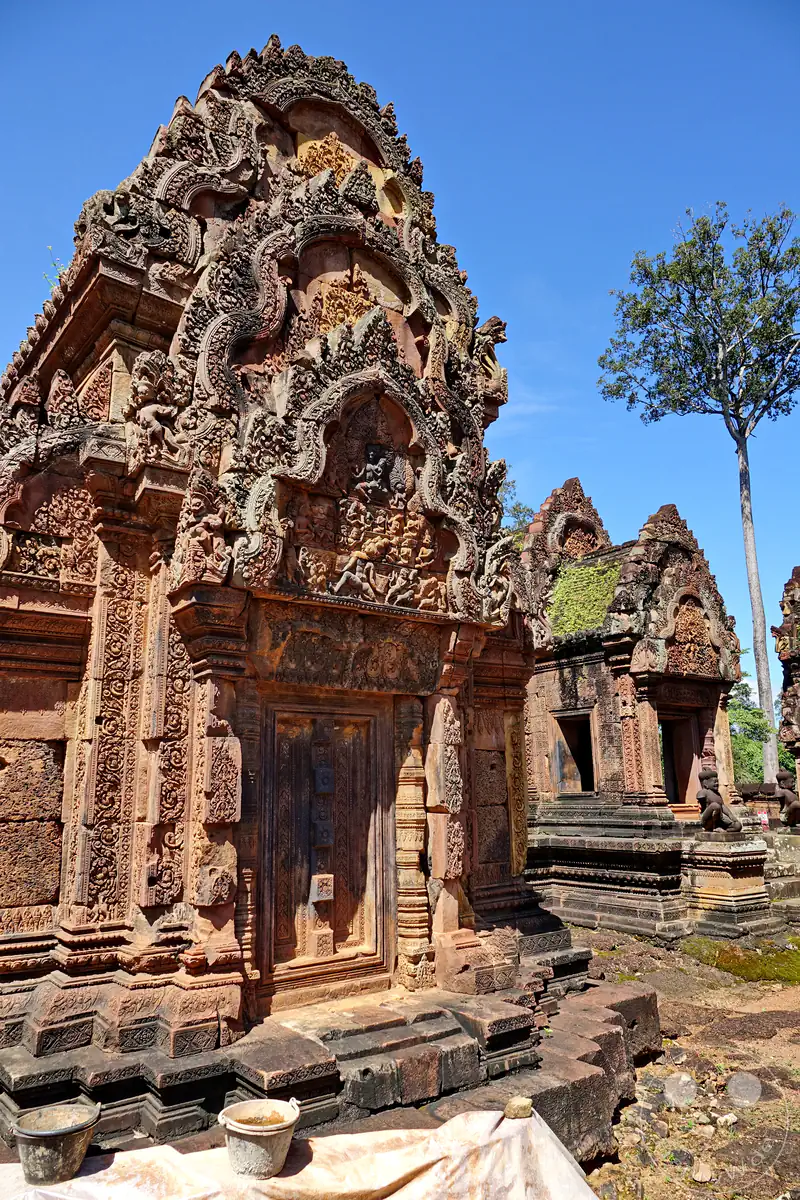 Eine detailreiche Nahansicht der monumentalen, reich verzierten Steintürme des Banteay-Srei-Tempels aus rotem Sandstein mit kunstvollen Giebelreliefs und Schnitzereien vor strahlend blauem Himmel.