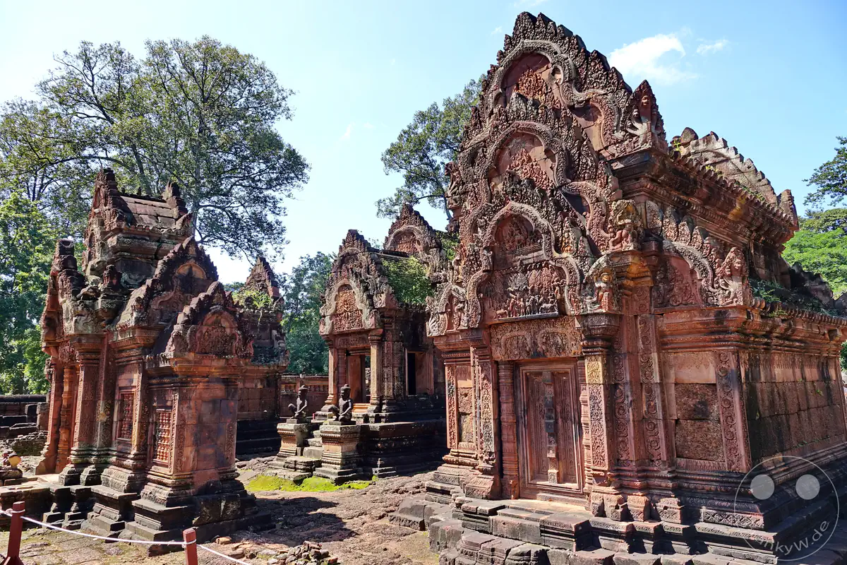 Die perspektivische Innenansicht des Banteay-Srei-Tempels aus rotem Sandstein mit seinen reich verzierten Steintürmen, kunstvoll geschichteten Giebelreliefs und steinernen Wächterfiguren im Hintergrund unter blauem Himmel.