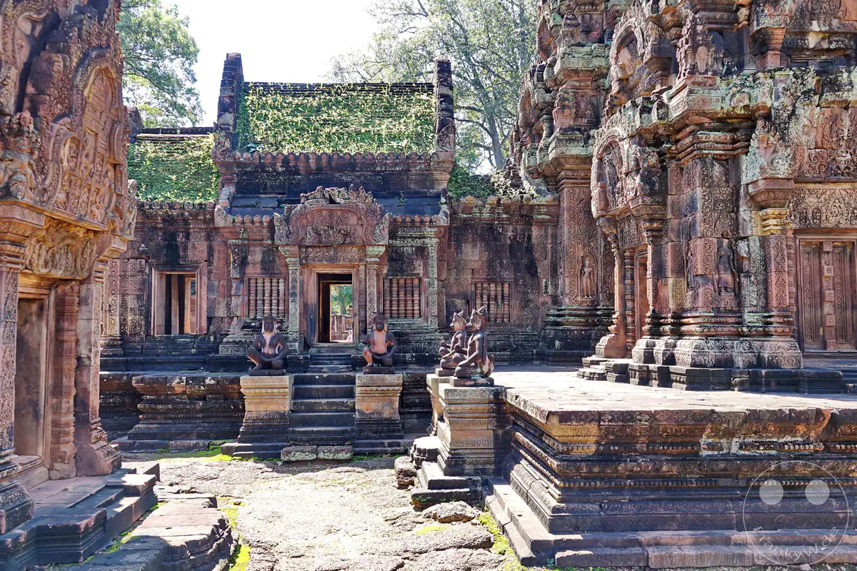 Der historische Innenhof des Banteay-Srei-Tempels aus rotem Sandstein mit moosbewachsenen Dächern, kunstvoll verzierten Steinfassaden und mehreren affenköpfigen Wächterstatuen auf erhöhten Plattformen.