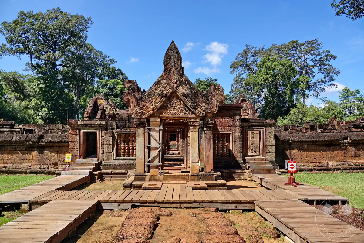 Die symmetrische Vorderansicht eines reich verzierten Steintors im Banteay-Srei-Tempel aus rotem Sandstein mit weitläufigen Holzstegen für Besucher, flankiert von historischen Umfassungsmauern und hohen Bäumen vor blauem Himmel.