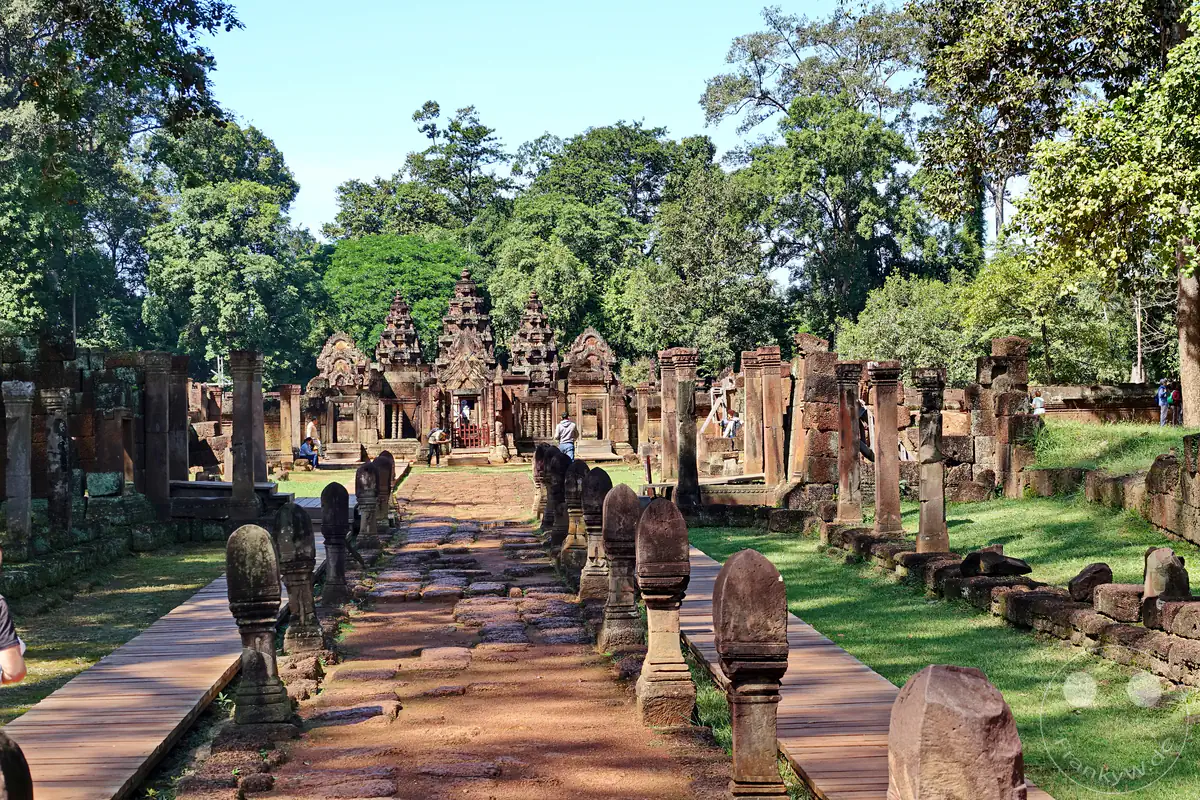 Der Blick entlang des zentralen, von steinernen Grenzpfosten gesäumten Prozessionsweges auf die drei markanten, reich verzierten Haupttürme des Banteay-Srei-Tempels inmitten grüner Bäume im Sonnenlicht.