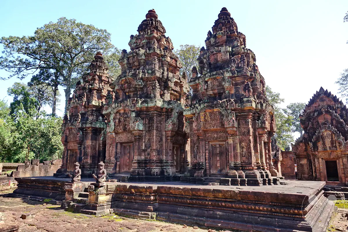 Die drei zentralen, monumentalen Steintürme des Banteay-Srei-Tempels aus rotem Sandstein auf einer erhöhten Plattform, flankiert von sitzenden Wächterstatuen und umgeben von grünen Bäumen unter klarem Himmel.