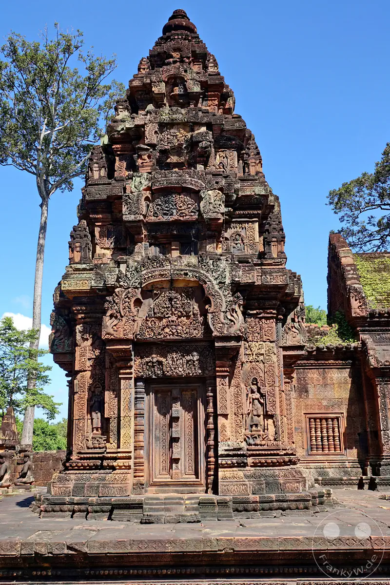 Die frontale Nahansicht eines reich verzierten Steinturms aus rotem Sandstein im Banteay-Srei-Tempel mit kunstvollen Giebelreliefs, Scheintüren und filigranen Devata-Skulpturen in den Mauernischen vor blauem Himmel.
