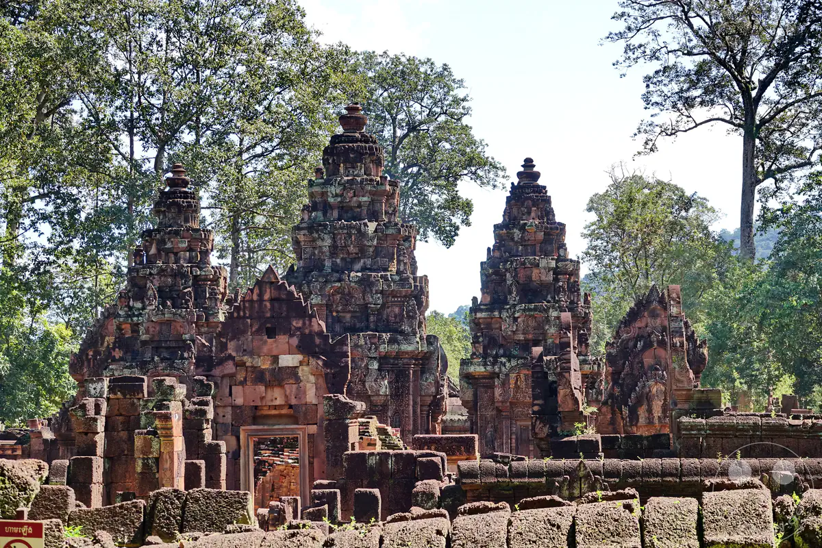 Die drei charakteristischen, reich verzierten Haupttürme des Banteay-Srei-Tempels aus rotem Sandstein hinter den historischen Außenmauern aus Lateritstein, umgeben von hohen Bäumen unter hellem Himmel.