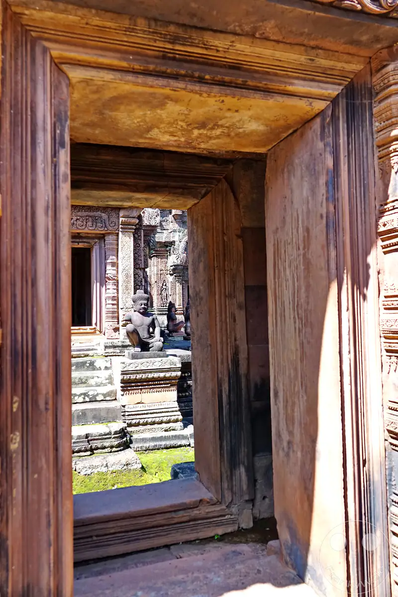 Der Blick durch den massiven Rahmen eines historischen Steintores im Banteay-Srei-Tempel auf eine sitzende, steinerne Wächterfigur im Innenhof der detailreich verzierten Anlage aus rotem Sandstein.