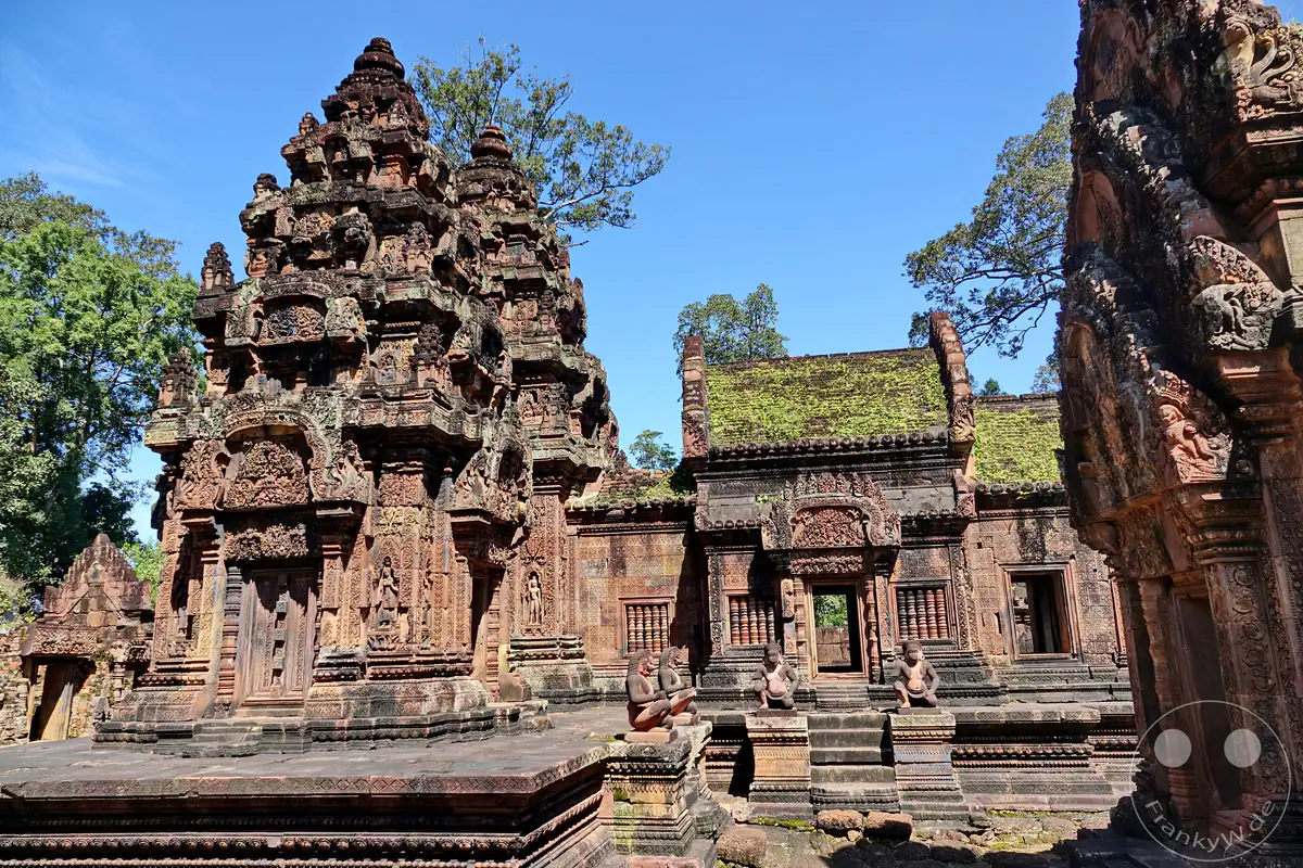 Die detailreiche Innenhofansicht des historischen Banteay-Srei-Tempels mit kunstvoll verzierten Steintürmen aus rotem Sandstein und affenköpfigen Wächterstatuen vor einem klaren blauen Himmel.
