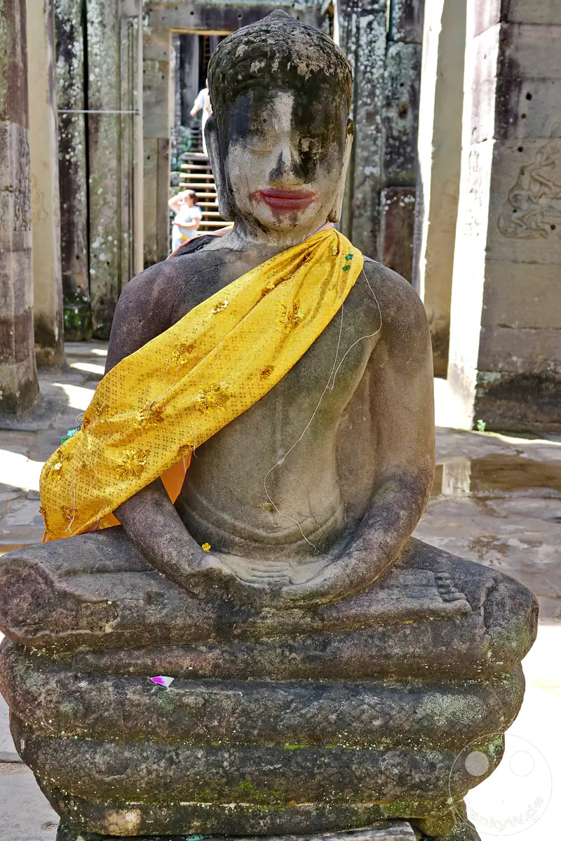 Eine historische, sitzende Buddha-Statue aus Stein im Bayon-Tempel in Kambodscha, geschmückt mit einer gelb-goldenen Schärpe inmitten der antiken Tempelruinen.