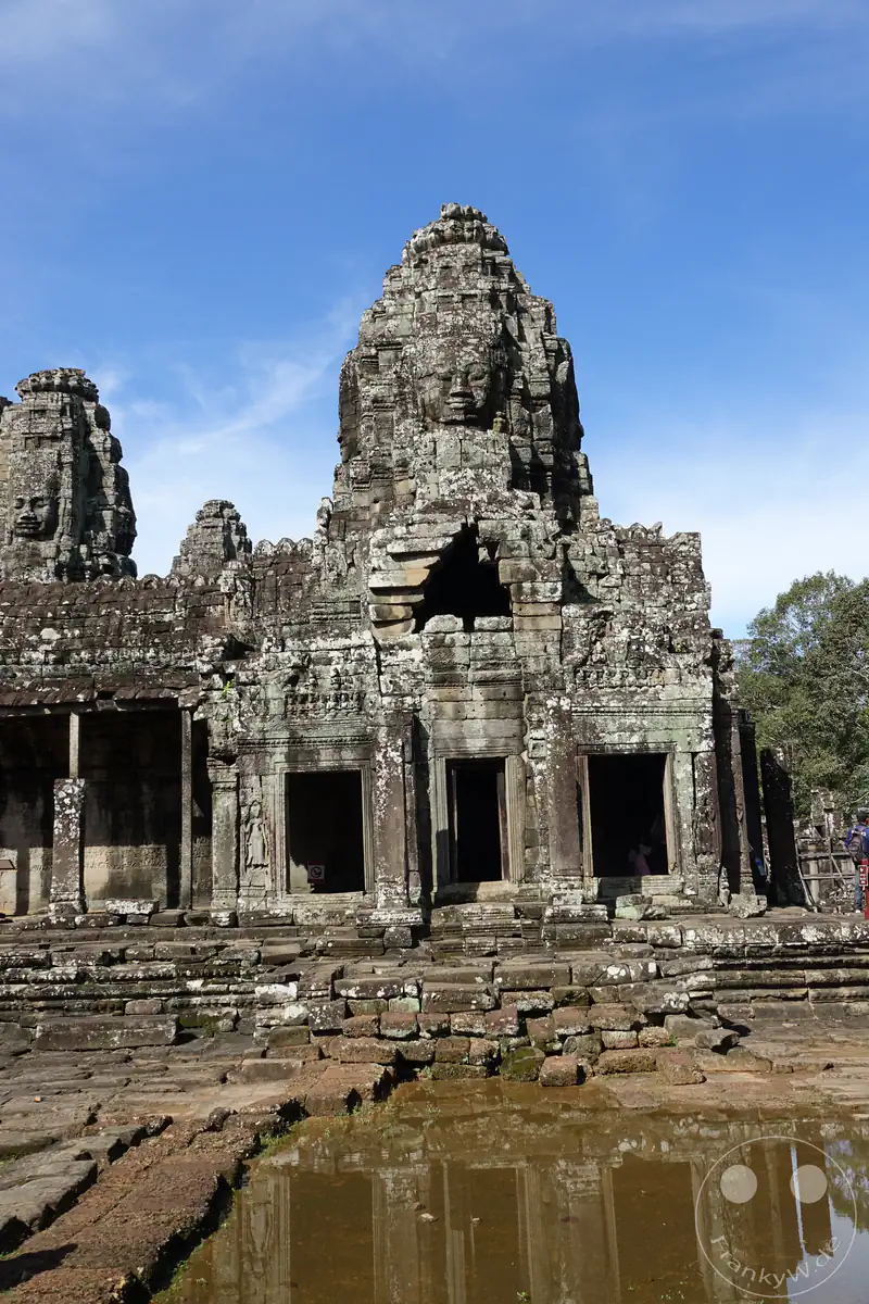 Die Spiegelung der historischen Steintürme mit den markanten Gesichtern des Bayon-Tempels in Angkor Thom, Kambodscha, in einer Wasserpfütze im Vordergrund an einem sonnigen Tag.