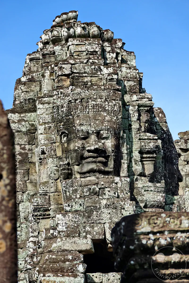 Nahaufnahme eines der lächelnden, monumentalen Steingesichter auf einem Turm des historischen Bayon-Tempels in Angkor Thom, Kambodscha, vor strahlend blauem Himmel.