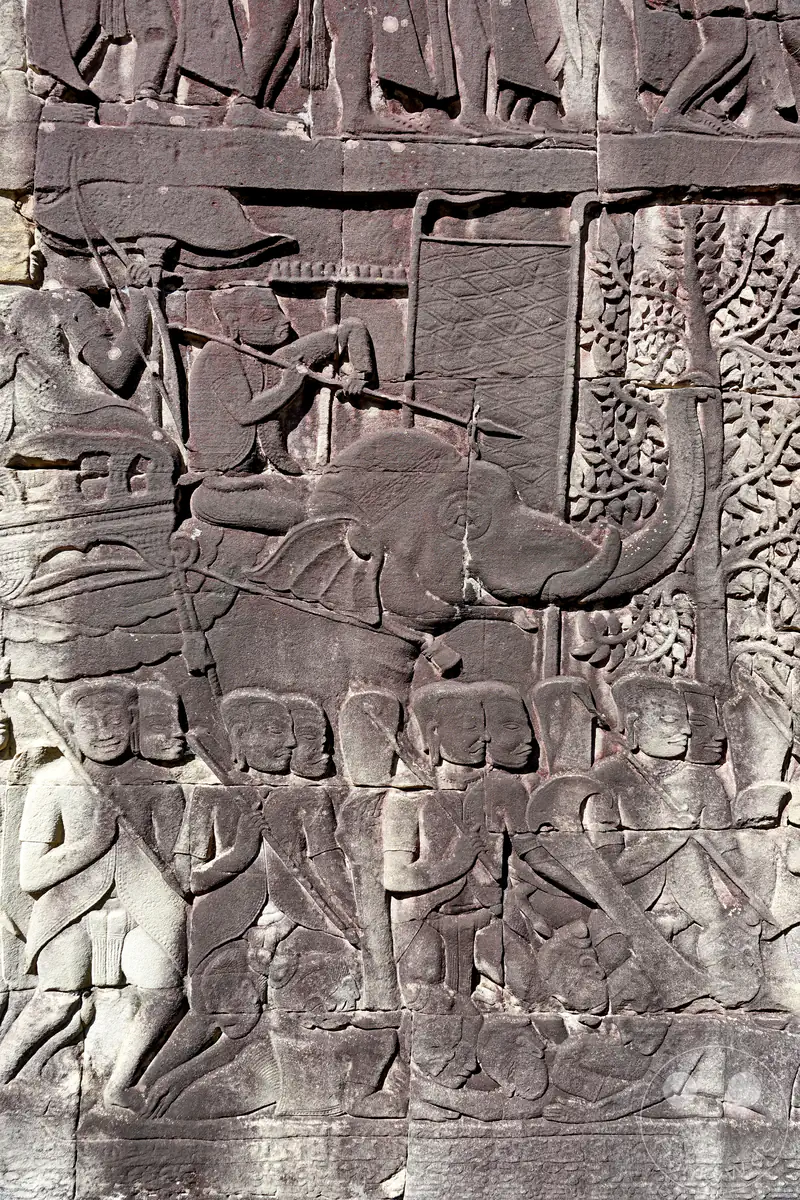 Ein historisches Steinrelief an einer Tempelmauer im Bayon-Tempel von Angkor Thom in Kambodscha, das Soldaten und einen Kriegselefanten in einer Marsch- oder Kampfszene darstellt