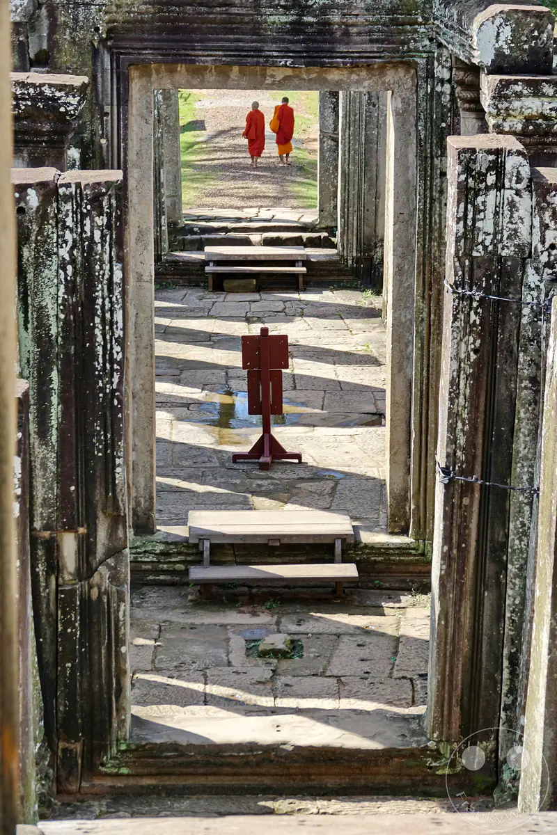 Der Blick durch eine Reihe historischer Steintore im Bayon-Tempel in Kambodscha, während im Hintergrund zwei buddhistische Mönche in orangefarbenen Roben laufen.