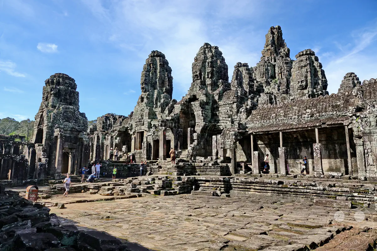 Die historischen Steintürme mit den markanten Gesichtern des Bayon-Tempels im Zentrum von Angkor Thom in Kambodscha mit Besuchern auf dem gepflasterten Innenhof unter blauem Himmel.
