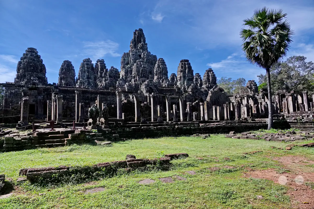 Die weite Außenansicht des historischen Bayon-Tempels in Angkor Thom mit seinen markanten, monumentalen Steintürmen hinter einer grünen Wiese und einer einzelnen Palme unter blauem Himmel.